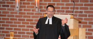 Ein Mann in einer schwarzen Robe steht an einer Kanzel und hält eine Predigt in einer Kirche. Die Kanzel ist mit drei Kerzen geschmückt, und die Wände bestehen aus roten Ziegelsteinen.