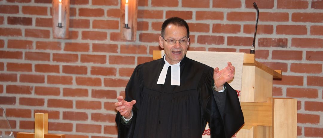 Ein Mann in einer schwarzen Robe steht an einer Kanzel und hält eine Predigt in einer Kirche. Die Kanzel ist mit drei Kerzen geschmückt, und die Wände bestehen aus roten Ziegelsteinen.