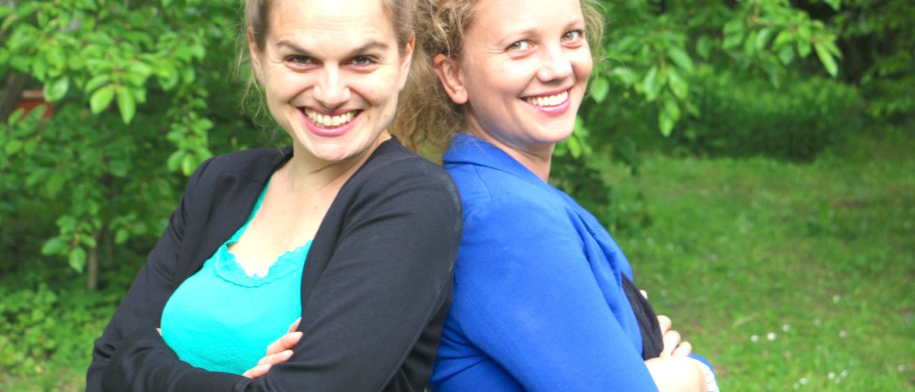 Zwei Frauen stehen eng beieinander, lächeln und posieren für ein Foto in einem Garten mit grünen Blättern und Gras.