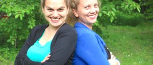 Zwei Frauen stehen eng beieinander, lächeln und posieren für ein Foto in einem Garten mit grünen Blättern und Gras.