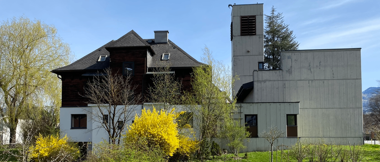 Ein großes braunes Haus mit schwarzem Dach, einem Schornstein und einem Turm auf der rechten Seite. Bäume umgeben das Haus und ein gelber Busch steht im Vordergrund.