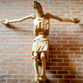 Ein hölzernes Kruzifix ist an einer Ziegelwand befestigt. Die Figur hat ausgestreckte Arme und scheint an der Wand genagelt zu sein.