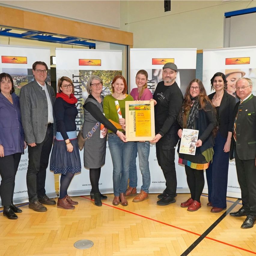 A group of people stands in front of banners, one holding a framed award. They all look at the camera. Behind them are two red lines on the floor.