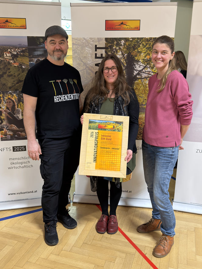 Three individuals stand indoors, one woman holding a framed certificate, while the man and another woman smile, with banners in the background.
