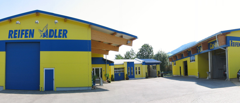 Ein großes gelbes und blaues Gebäude mit dem Namen Reifen Adler darauf. Es hat eine blaue Garagentür und ist von Bäumen umgeben.