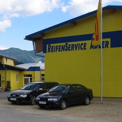 Ein gelbes und blaues Gebäude mit der Aufschrift Reifenservice Adler, vor dem zwei Autos geparkt sind und ein Fahnenmast mit einer Fahne.