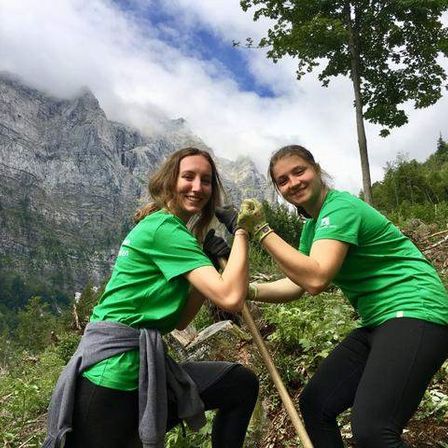 Zwei Frauen in grünen Shirts lächeln und posieren für ein Foto, während sie auf einem Pfad in einem Berggebiet arbeiten. Sie halten einen Stock.