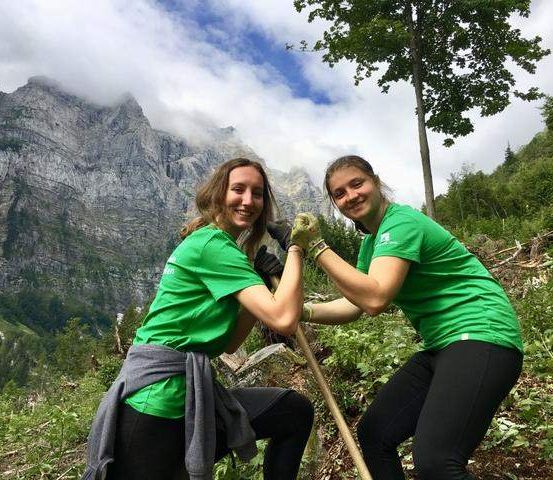 Zwei Frauen in grünen Shirts lächeln und posieren für ein Foto, während sie auf einem Pfad in einem Berggebiet arbeiten. Sie halten einen Stock.