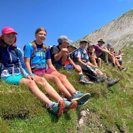 Eine Gruppe junger Wanderer mit Rucksäcken sitzt auf dem grasbewachsenen Hang eines Berges unter einem klaren blauen Himmel.