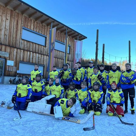 Eine Gruppe junger Eishockeyspieler in gelben und blauen Uniformen posiert für ein Foto auf einer Eisbahn vor einem Holzgebäude.
