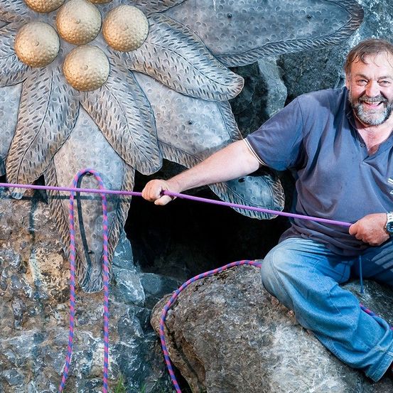Ein Mann in einem blauen Hemd und Jeans sitzt auf einem Felsen, hält eine lilafarbene Seil und lächelt. Er scheint an einer Aktivität beteiligt zu sein, möglicherweise Klettern.