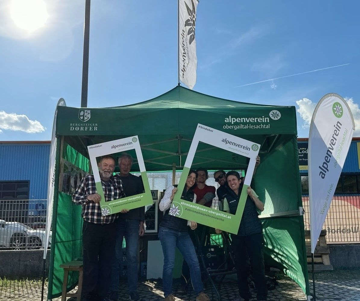 A group of people stand under a green tent, holding photo frames with the word 'alpenverein' written on them. The tent has the text 'BERGSTEIGER DORFER' and 'alpenverein obergailtal-lesachtal'. A flag is placed behind the tent.