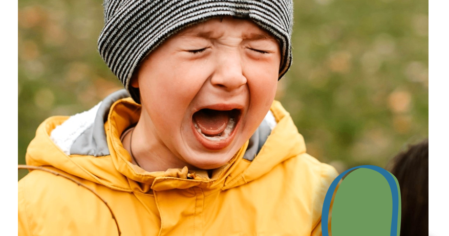 Ein Kind in einer gelben Jacke und einer grauen Mütze weint im Freien mit Gras im Hintergrund.