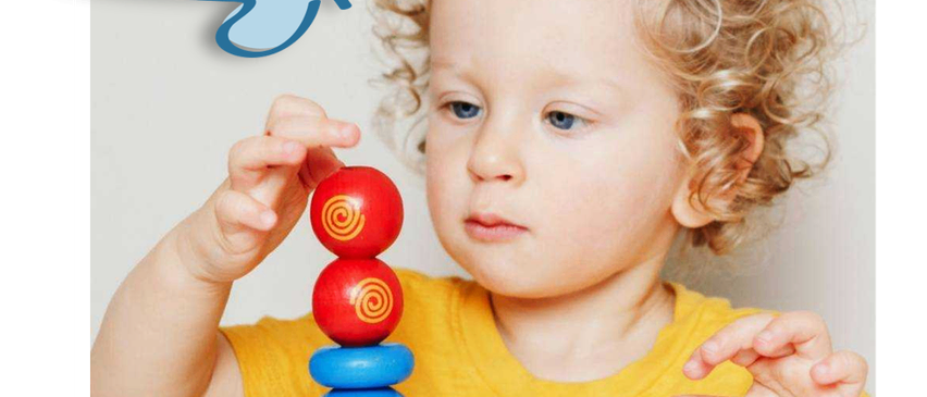 Ein kleines Kind mit lockigem Haar spielt mit einem bunten Stapel Ringe und hält einen roten Ring oben.
