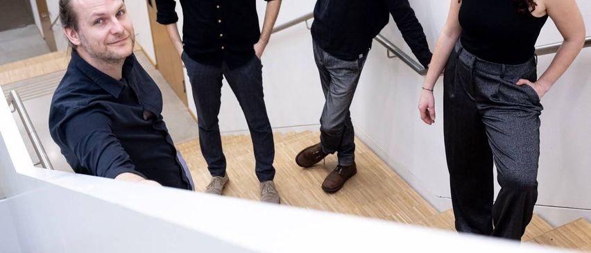 Vier Personen stehen auf den Treppen eines Gebäudes. Sie tragen alle schwarze Langarm-Shirts. Die Frau auf der rechten Seite hat lockige Haare und lächelt. Der Mann auf der linken Seite schaut zur Kamera.