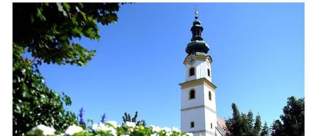 Ein hoher weißer Turm mit einer Uhr an der Seite steht in einem Park mit grünen Bäumen und einem klaren blauen Himmel.