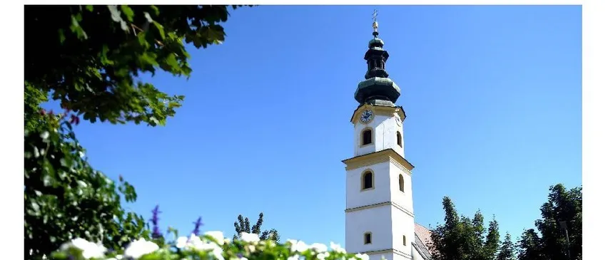 Ein hoher weißer Turm mit einer Uhr an der Seite steht in einem Park mit grünen Bäumen und einem klaren blauen Himmel.
