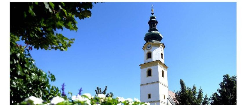 Ein hoher weißer Turm mit einer Uhr an der Seite steht in einem Park mit grünen Bäumen und einem klaren blauen Himmel.