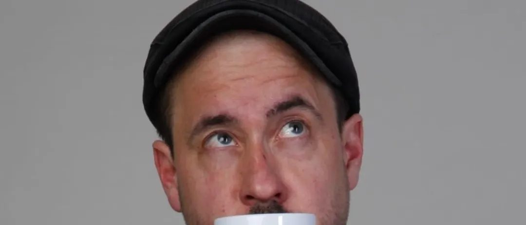 Ein Mann mit einer schwarzen Kappe trinkt aus einer weißen Tasse mit den Worten 'Beste Tante der Welt' darauf. Der Hintergrund ist grau.