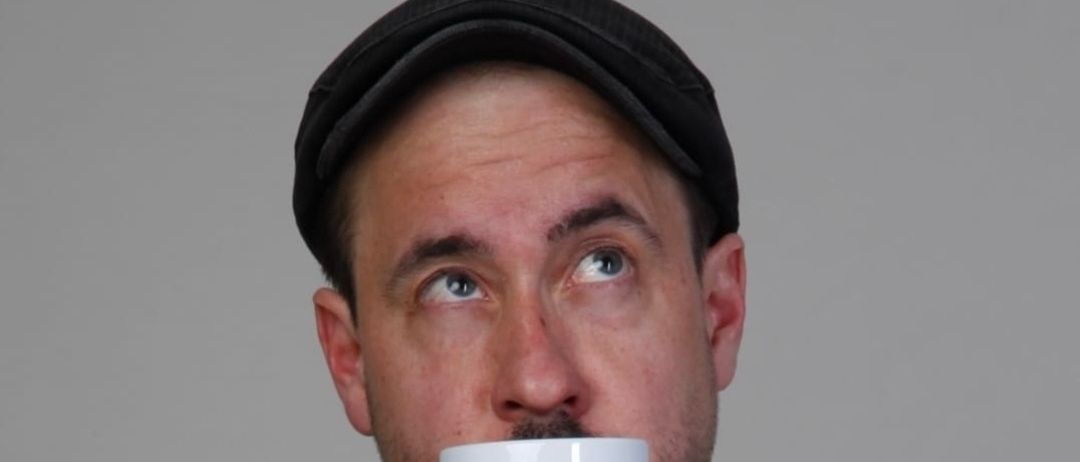 Ein Mann mit einer schwarzen Kappe trinkt aus einer weißen Tasse mit den Worten 'Beste Tante der Welt' darauf. Der Hintergrund ist grau.