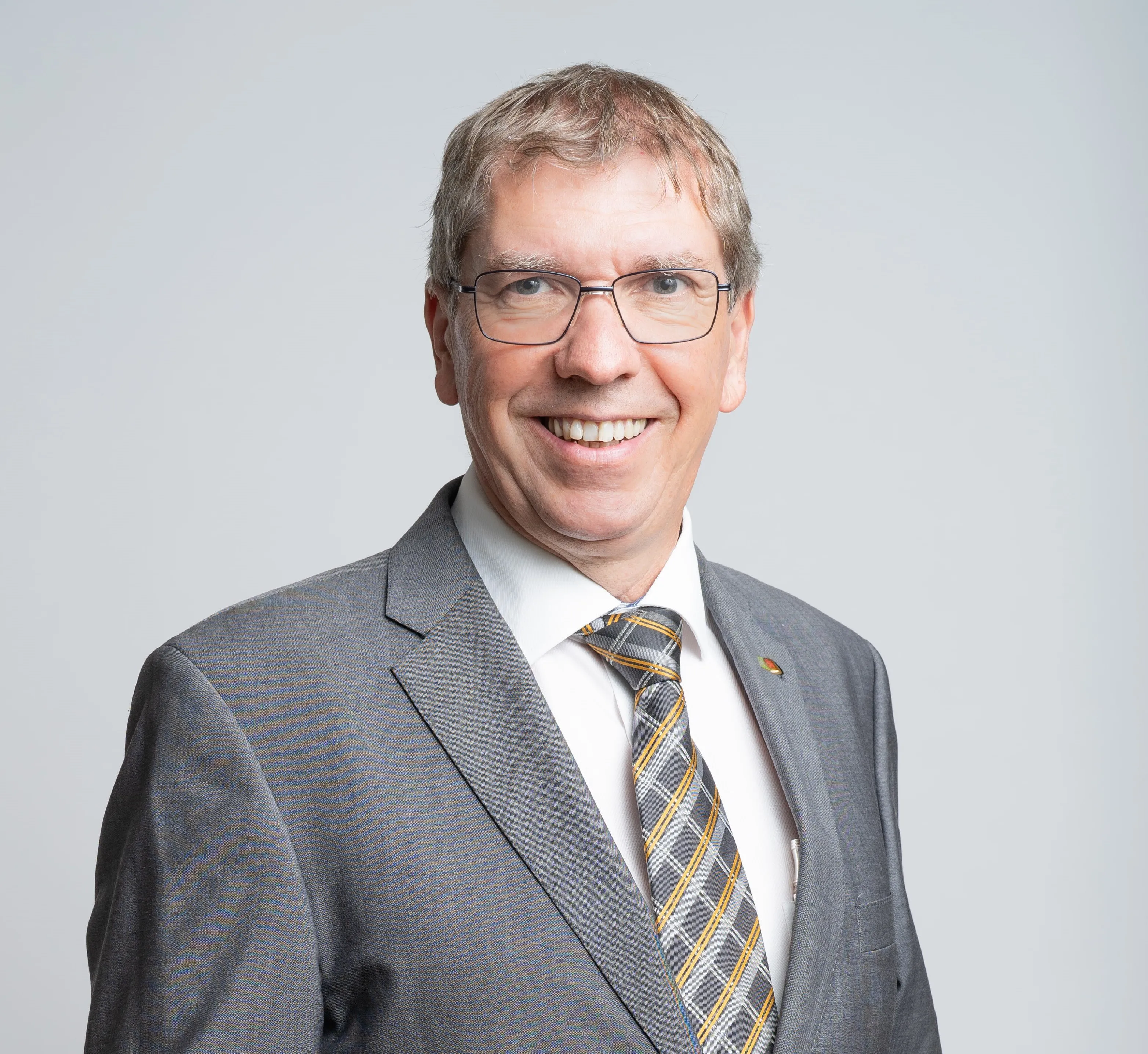 Ein Mann in formeller Kleidung, lächelt in die Kamera und trägt eine Brille und eine Krawatte.