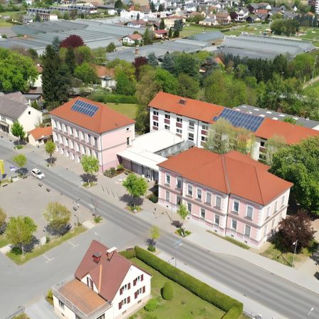 Eine Luftaufnahme eines Wohngebiets mit Häusern, einer Straße und Solarmodulen auf den Dächern. Bäume und Büsche umgeben das Gebiet. Ein Auto ist am Straßenrand geparkt.
