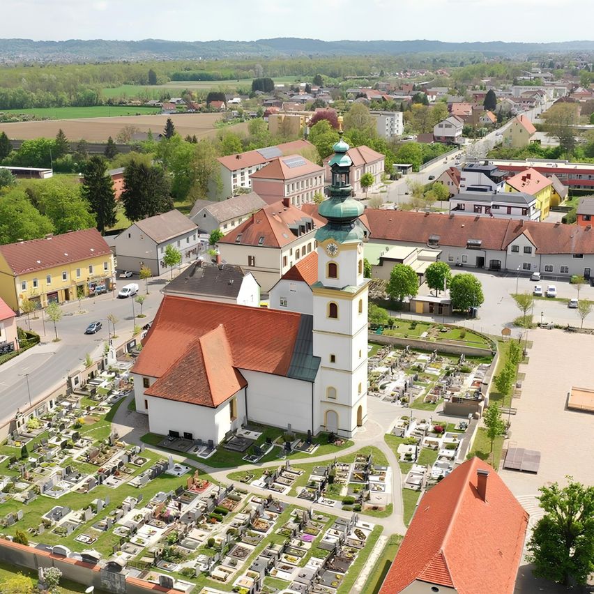 Luftaufnahme einer Stadt mit einer Kirche, einem Friedhof, Häusern und grünen Feldern. Die Stadt hat viele Gebäude, einige mit Solarmodulen.