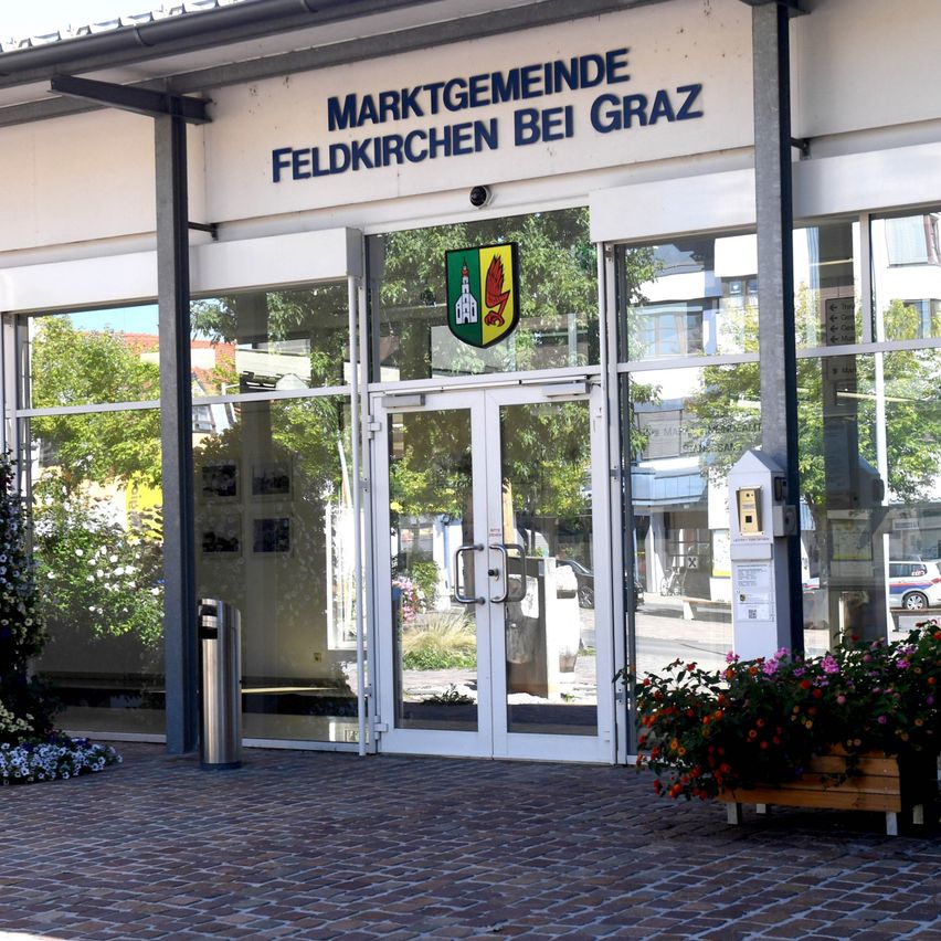 Der Eingang von Marktgemeinde Feldkirchen bei Graz hat eine Glastür mit Logo, Blumen und einem Mülleimer.