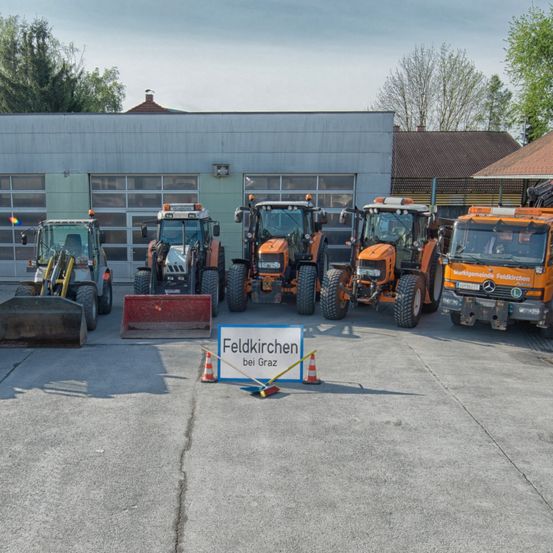 Mehrere orange Traktoren und ein gelber Bagger sind vor einem Gebäude mit einem Schild, das Feldkirchen bei Graz steht, geparkt.