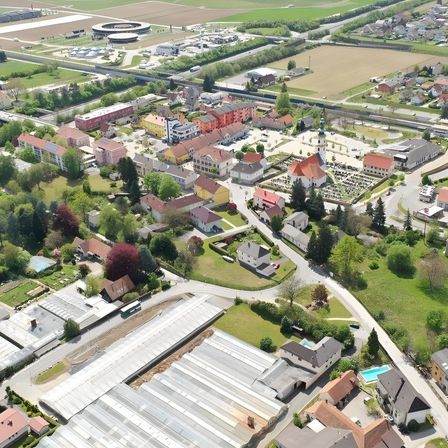 Eine Luftaufnahme einer Stadt mit vielen Gebäuden, Straßen und viel Grün. Die Stadt ist von Bäumen umgeben und hat eine Mischung aus Wohn- und Geschäftsgebäuden. In der Ferne befindet sich ein großes Feld mit einigen Strukturen und einem runden Gebäude.