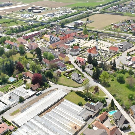 Eine Luftaufnahme einer Stadt mit vielen Gebäuden, Straßen und viel Grün. Die Stadt ist von Bäumen umgeben und hat eine Mischung aus Wohn- und Geschäftsgebäuden. In der Ferne befindet sich ein großes Feld mit einigen Strukturen und einem runden Gebäude.
