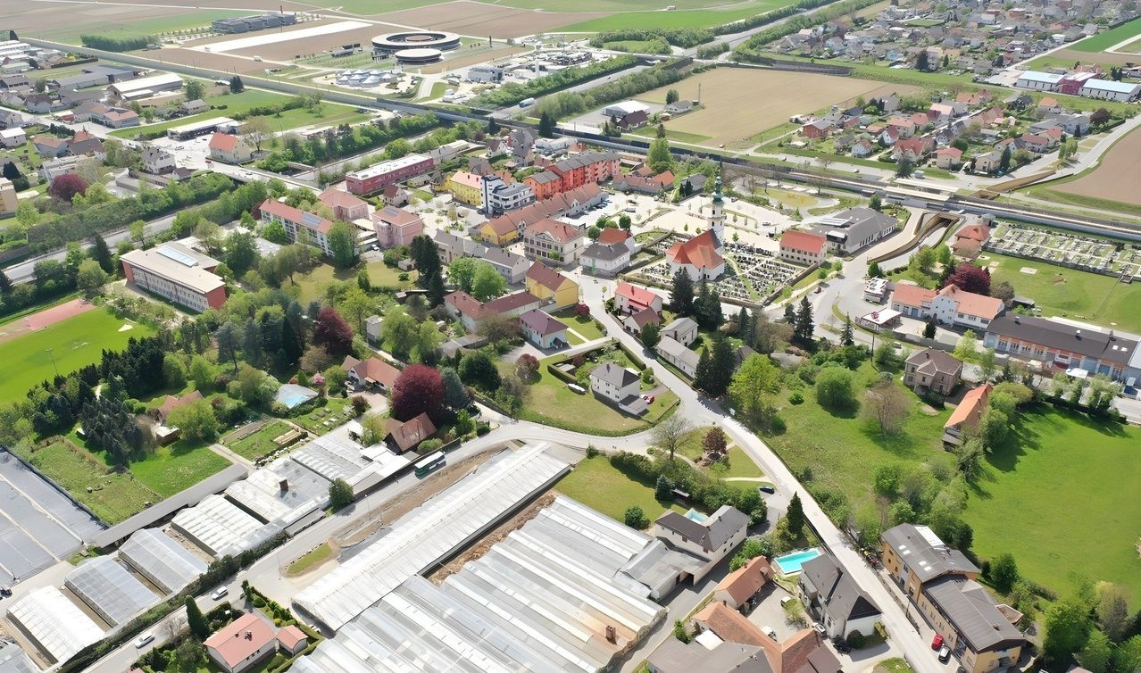 Eine Luftaufnahme einer Stadt mit vielen Gebäuden, Straßen und viel Grün. Die Stadt ist von Bäumen umgeben und hat eine Mischung aus Wohn- und Geschäftsgebäuden. In der Ferne befindet sich ein großes Feld mit einigen Strukturen und einem runden Gebäude.