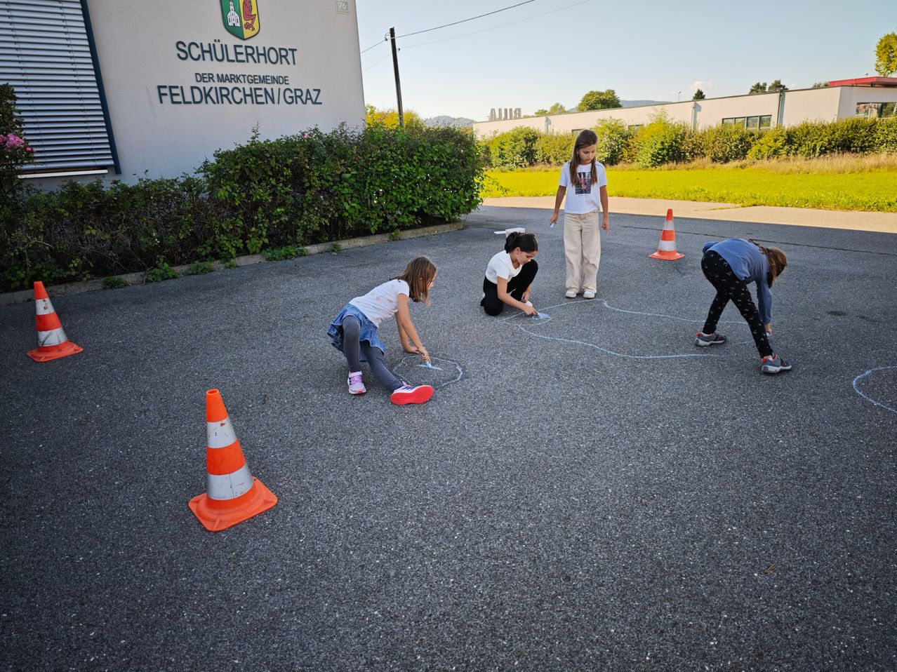 Vier Mädchen spielen mit Kreide auf einer gepflasterten Fläche mit orangefarbenen Verkehrskegel. Das Gebäude hinter ihnen trägt die Aufschrift 'Schulerhort der Marktgemeinde Feldkirchen/Graz'.