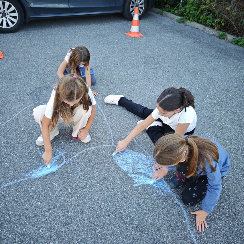 Vier Mädchen zeichnen mit Kreide auf dem Bürgersteig. Sie sind von einem Verkehrskegel und einem blauen Auto umgeben.