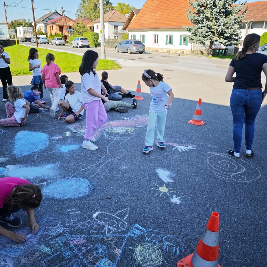 Eine Gruppe von Kindern malt mit Kreide auf dem Bürgersteig. Sie sind von Verkehrskegel umgeben. Eine erwachsene Frau beobachtet von der Seite.