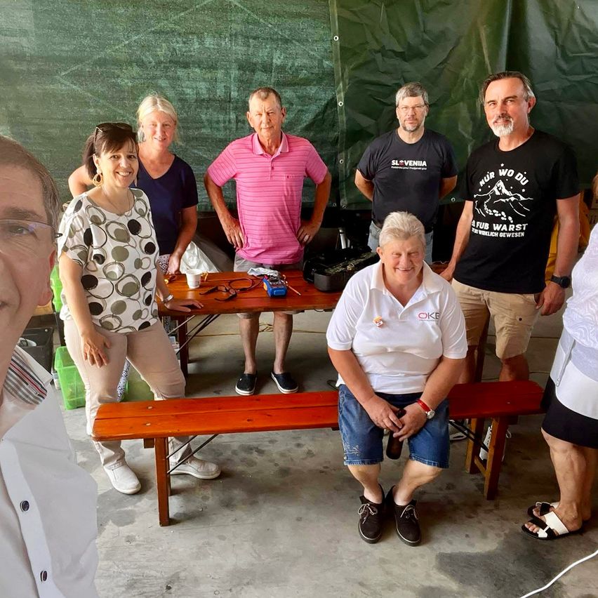 Eine Gruppe von Menschen steht und sitzt um einen Tisch in einem Raum. Eine Person sitzt auf einer Bank. Eine Frau in einem gepunkteten Shirt steht neben dem Tisch.