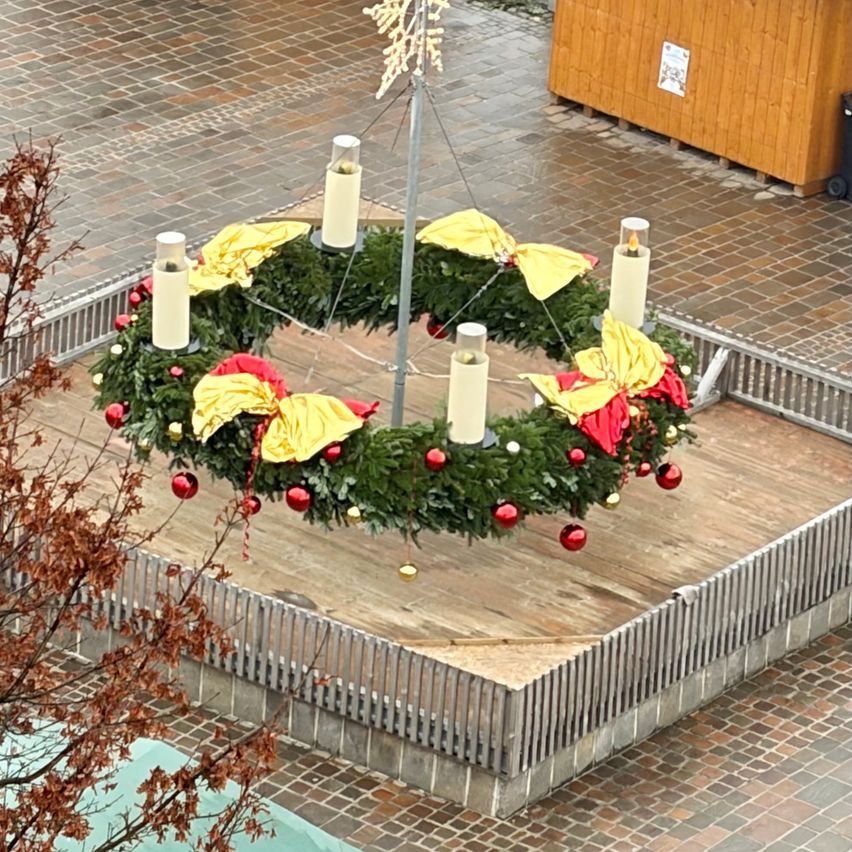 Ein kreisförmiger Kranz wird auf einer hölzernen Plattform mit einer silbernen Brüstung ausgestellt. Er hat gelbe Bänder, rote Ornamente und weiße Kerzen. Der Kranz ist von einem Ziegelboden umgeben.