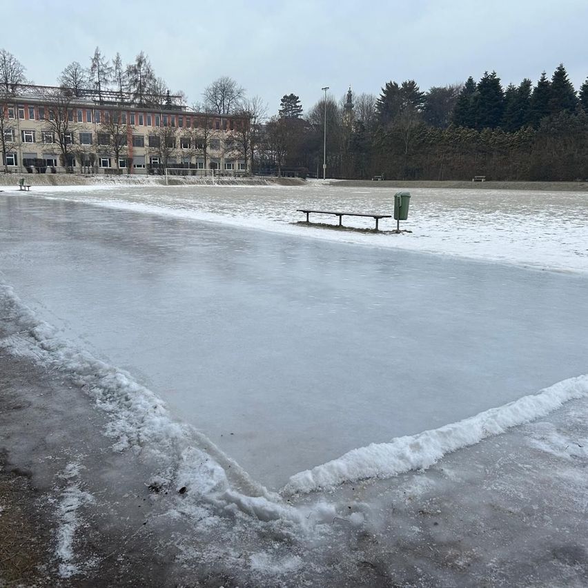 Eine leere Eisbahn mit einer Bank, einem Mülleimer und einem großen Gebäude mit mehreren Fenstern im Hintergrund. Der Boden ist mit Schnee bedeckt.