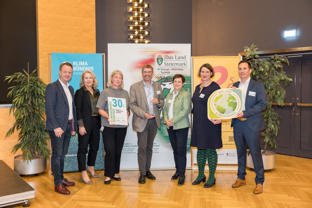 Eine Gruppe von Menschen steht zusammen und hält Auszeichnungen und Schilder. Einer hält ein Schild mit 'Klima Bündnis' und ein anderer ein Schild mit einem Globus. Im Hintergrund sind Banner und Lampen zu sehen.