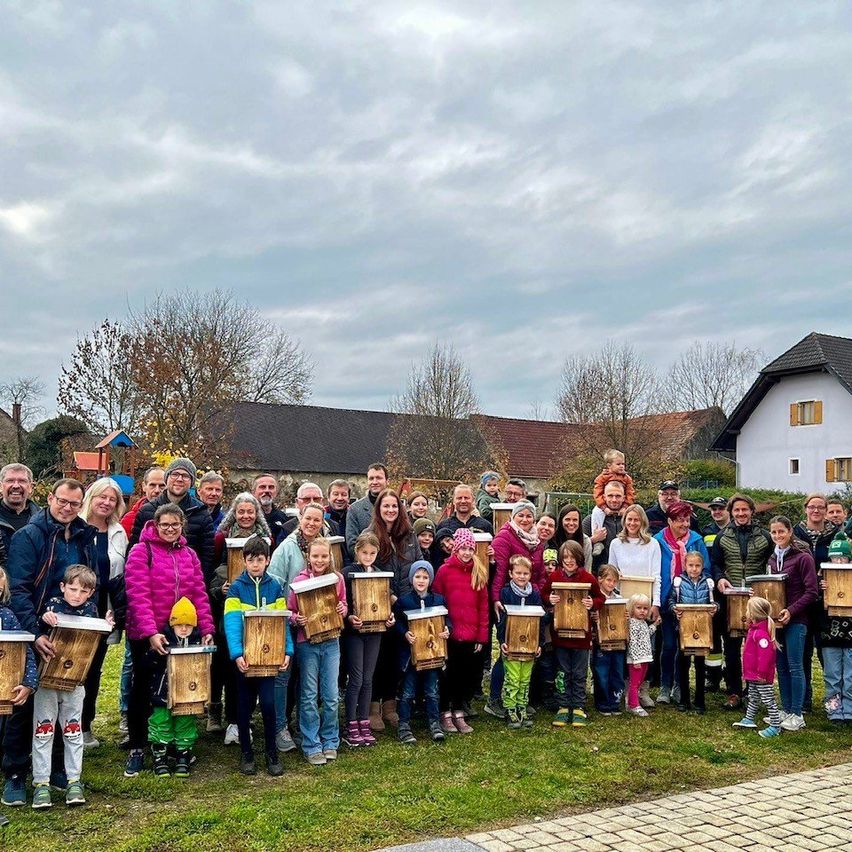 Eine Gruppe von Menschen posiert für ein Foto im Park, wobei sie Holzkisten halten. Der Hintergrund besteht aus Häusern und Bäumen unter einem bewölkten Himmel.