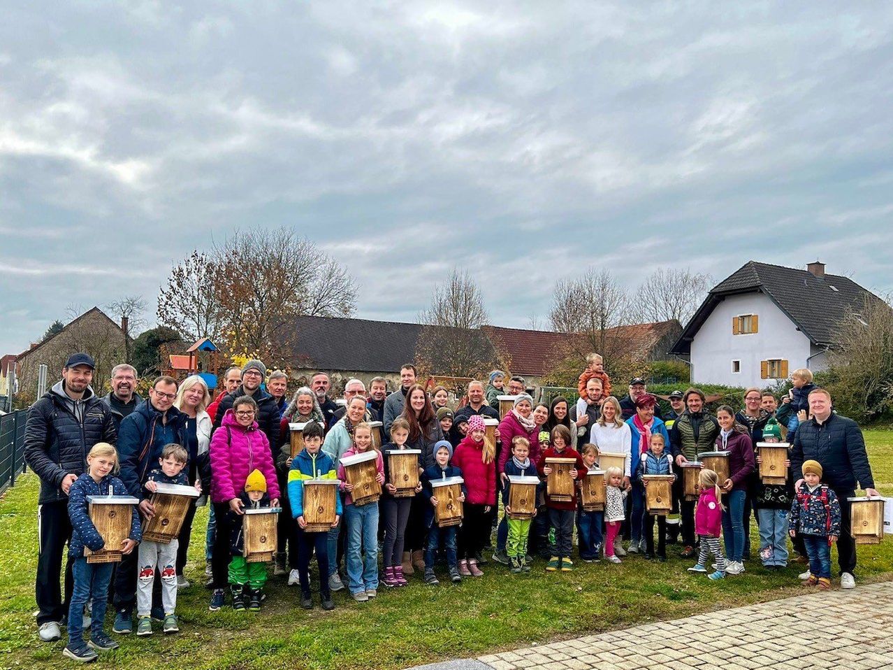 A group of people are posing for a photo in a park, holding wooden boxes. The scene is set against a backdrop of houses and trees under a cloudy sky.