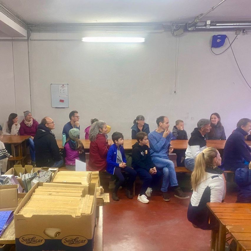 Ein Raum mit weißen Wänden, einer Deckenleuchte und einer Tafel. Mehrere Personen sitzen auf Bänken, einige schauen zu einer Frau, die vor ihnen steht. Im Vordergrund sind Kisten gestapelt.