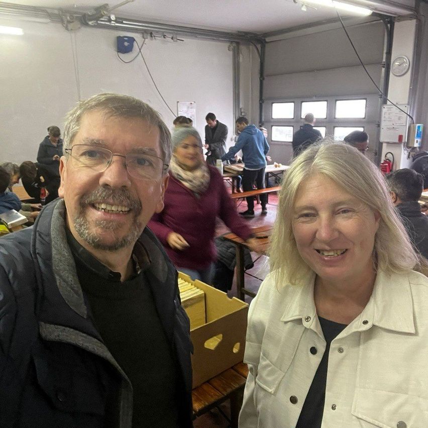 A man and a woman smile for a selfie in a room with several people. Behind them, a woman holds a box. Tables, benches, and a clock are visible.