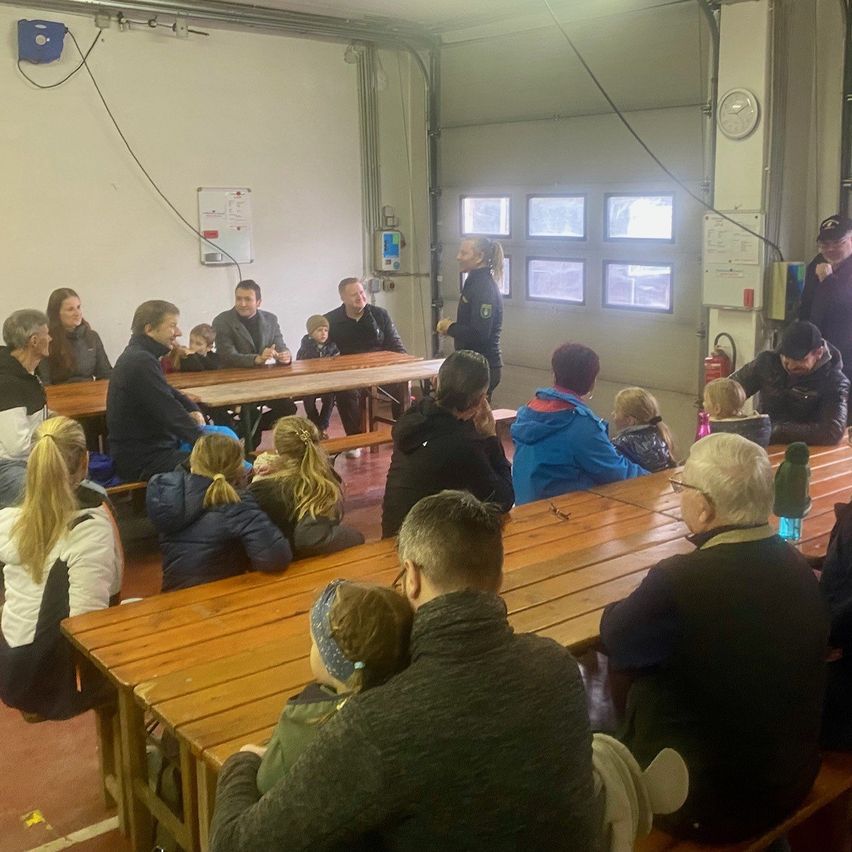 A group of people sits around long tables in a room with white walls. A woman stands in front of them, perhaps giving a presentation. There are children sitting on the tables, and a fire extinguisher is mounted on the wall.
