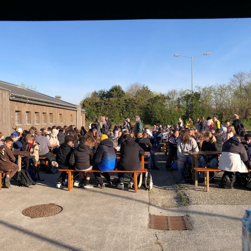 Eine Gruppe von Menschen sitzt auf Holzbänken im Freien in der Nähe eines Gebäudes unter einem klaren Himmel, wobei eine Straßenlampe im Hintergrund zu sehen ist.