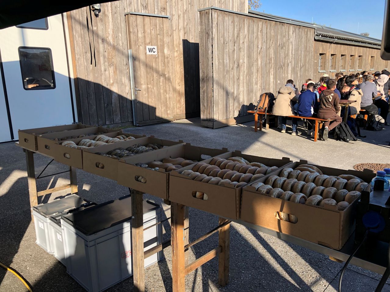 Ein Holzgebäude mit WC-Schild hat zwei Türen. Leute sitzen auf einer Bank davor. Ein Tisch hält Kartons mit Bagels.