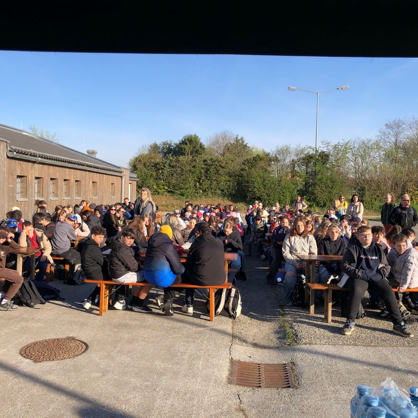 Eine große Gruppe von Menschen, möglicherweise Schüler, sitzt auf Bänken im Freien. Sie tragen verschiedene Jacken und Mützen. Flaschen liegen auf dem Boden, und ein großes Gebäude ist im Hintergrund.