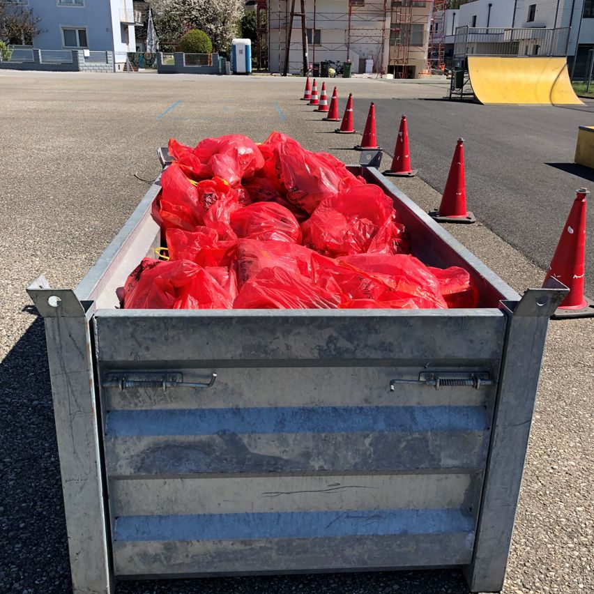 Ein mit roten Plastiktüten gefüllter Metallcontainer ist von orangefarbenen Verkehrskegel umgeben auf einem gepflasterten Bereich. Gebäude und eine Rampe sind im Hintergrund.