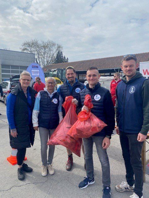 Fünf Personen, wahrscheinlich Freiwillige, stehen in einem Außenbereich und halten rote Tüten in den Händen. Hinter ihnen befindet sich ein Gebäude mit einem Banner und einem geparkten Fahrzeug.