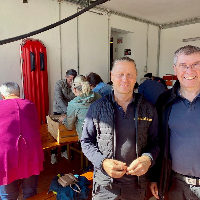 Eine Gruppe von Menschen ist drinnen, zwei Männer stehen eng beieinander und lächeln. Eine Frau in einem rosa Pullover schaut etwas im Hintergrund an.